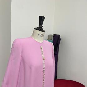 "Vybrali jsme lehký vlněný žoržet v pastelové růžové barvě. Ten nechal vyniknout velkorysé splývavé siluetě šatů s jemným tvarováním živůtku, úzkým rukávům s kulatými rameny, rolákovou vázačkou u krku a volnými objemnými zády s decentní vlečkou," říká o modelu návrhář Král.