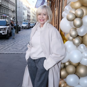 Jana Švandová obléká béžový podzimní kabát v kombinaci s šedými kalhotami s vysokým pasem a bílým rolákem. 